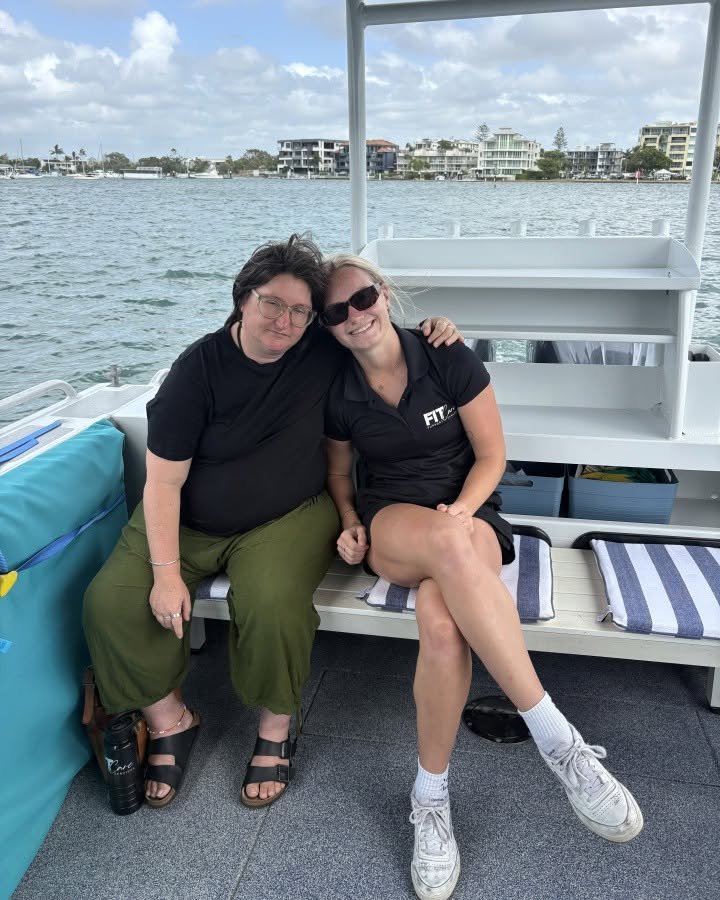 A FITCare support worker and participant sit arm in arm, smiling, on a boat cruising calm coastal waters with waterfront homes behind them