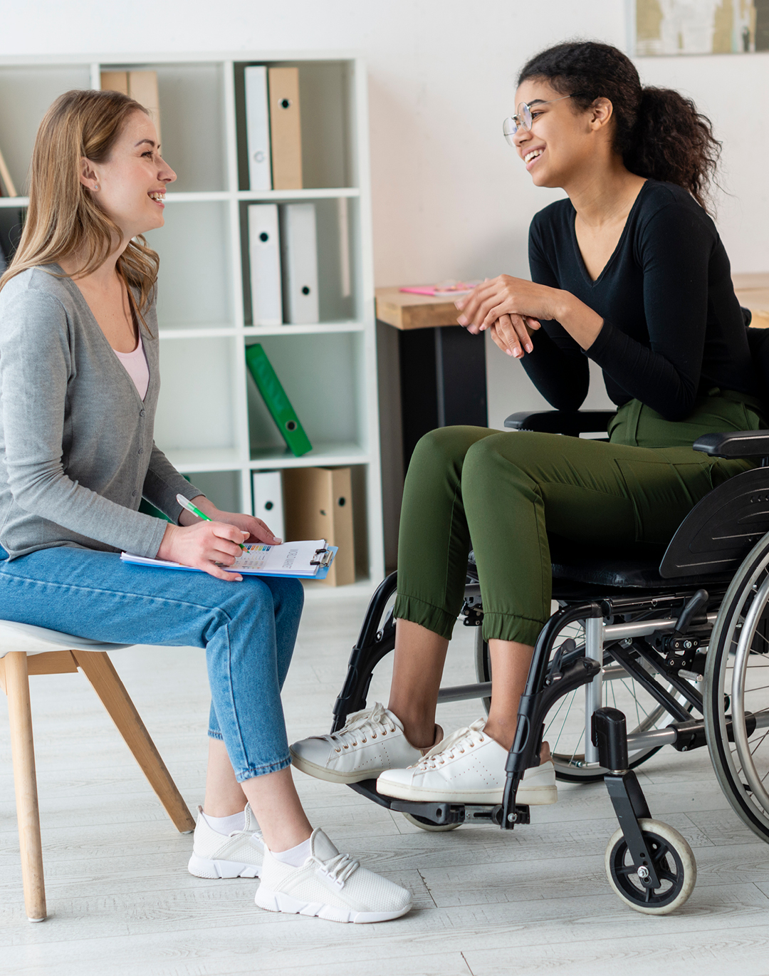 A FITCare support worker and participant sit together smiling during a catch-up, clipboard in hand