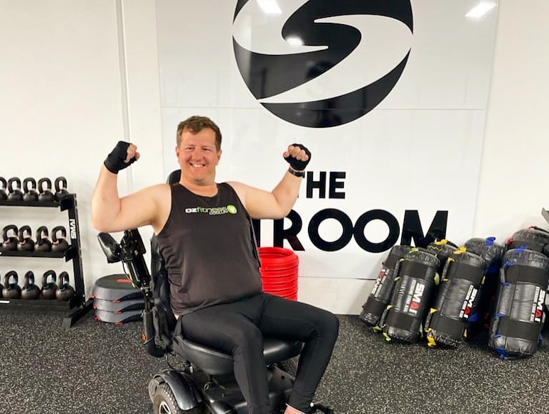 A FITCare participant in a wheelchair flexes after a gym session, smiling in a fitness studio
